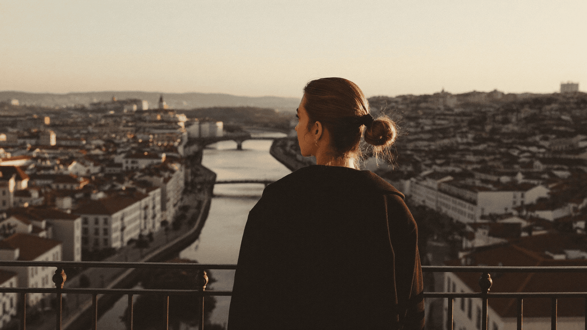Person at a Lisbon miradouro railing at golden hour, looking out over rooftops and the river